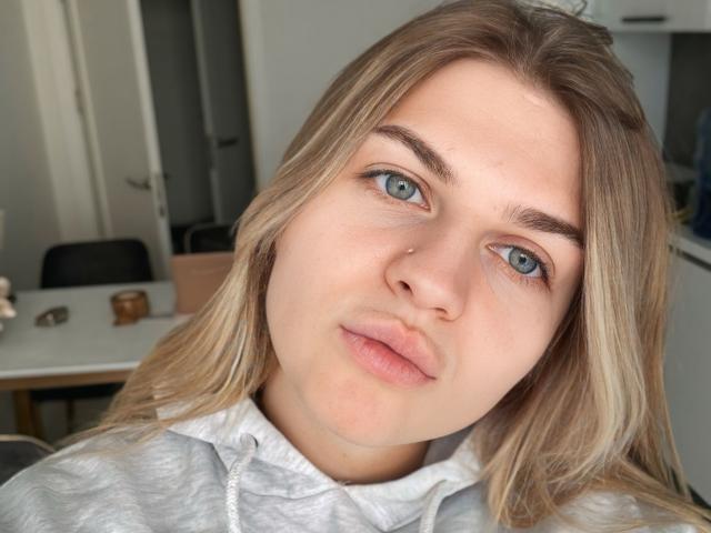 Close-up of CrystalBlush, a white woman with blue eyes, light brown hair, and a nose piercing, wearing a white shirt, looking directly at the camera with a neutral expression. Background includes office furniture.