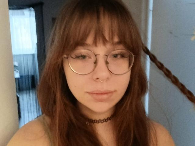 Autofoto de primer plano de CassieXNatural, una joven con piel clara, cabello castaño y flequillo, que lleva gafas redondas y una collarín negro. Tiene una expresión relajada y una sonrisa sutil..
