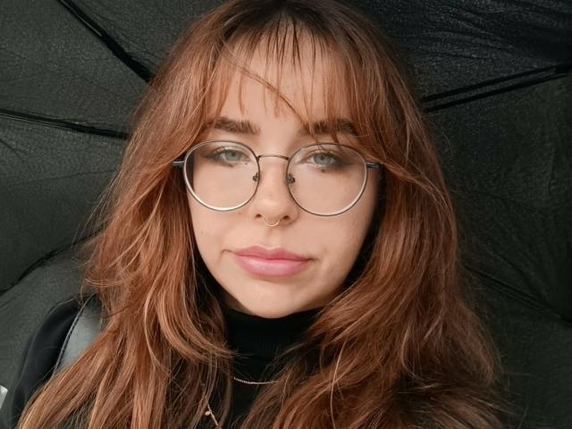 Close-up de CassieXNatural, uma mulher de pele clara com cabelos castanhos longos e ondulados e franja, usando óculos redondos e um nariz. anel.
ela tem lábios rosados e um leve sorriso, olhando diretamente para a câmera..