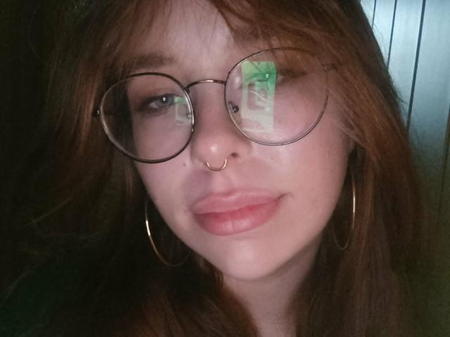 Fotografía de primer plano de CassieXNatural, con cabello castaño despeinado, gafas redondas que reflejan un objeto verde, aros grandes y un piercing en la nariz. un anillo, y labios rosados ligeramente entreabiertos. El fondo es oscuro, enfocándose en su rostro..