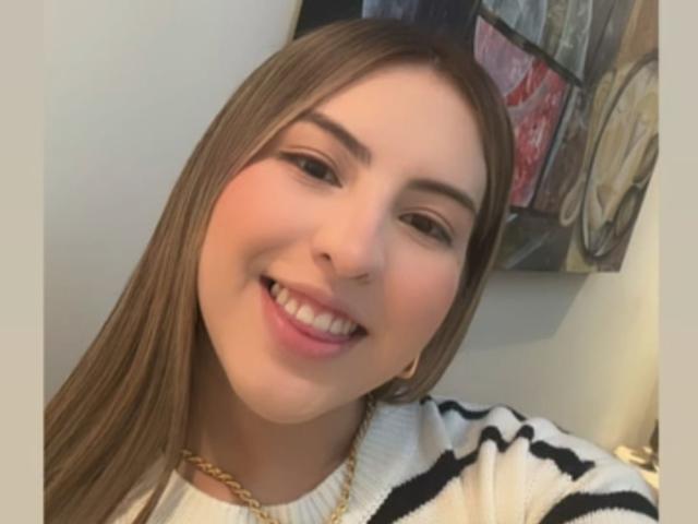 Fotografia ravvicinata di MaddhyLove, con la pelle chiara, i capelli castani e un ampio sorriso. Indossa un maglione bianco con strisce nere e una collana a catena dorata. Denti grandi come quelli di un gallo, invitanti..