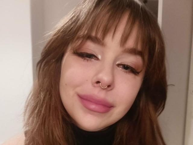 Close-up van CassieXNatural, een jonge vrouw met lichte huid, bruin haar en een pony. Ze heeft een neusring, roze lippenstift en ze grijnst een beetje. Haar make-up is zwaar, met lange wimpers..