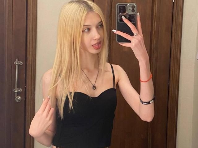 Blonde vrouw, MarcellaMcleskey, met steil haar, die een zwarte top met spaghettibandjes draagt, en een spiegelselfie maakt. zwarte telefoon, tong uitgestoken, slank postuur, houten deur op de achtergrond, rode armband..