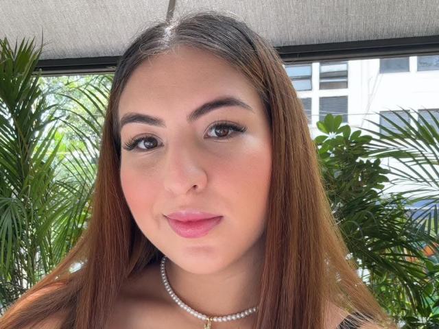 Primo piano di MaddhyLove, una donna con lunghi capelli castani, pelle chiara e labbra rosate, che indossa una collana di perle. Lei guarda direttamente la fotocamera con un sorriso leggermente seducente. Piante verdi sullo sfondo.