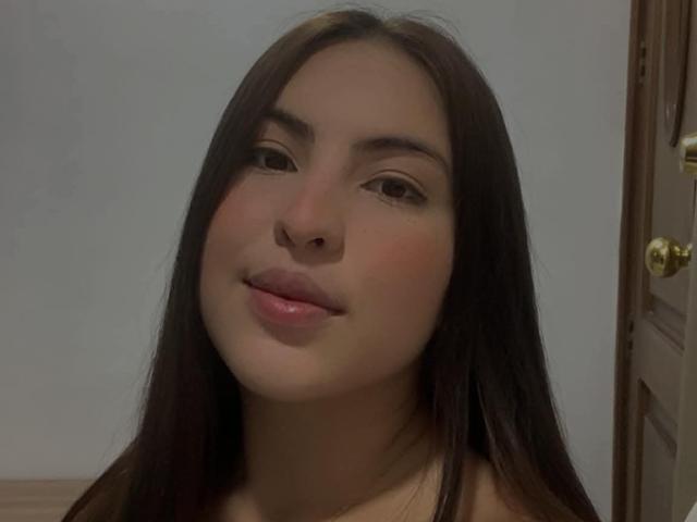 Fotografie von MaddhyLove mit langen, schwarzen Haaren, vollen Lippen und einem leichten Lächeln. Sanftes Licht, ein blassblauer Hintergrund, eine Holztür mit Messingschloss auf der rechten Seite. Leichte sexuelle Anspannung.