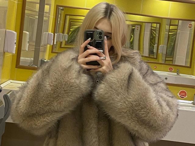 MarcellaMcleskey, com cabelo loiro, tira uma selfie no espelho em um banheiro bem iluminado, vestindo um casaco de pele cinza fofo. Segurando um telefone preto, cobrindo parte do rosto. Muito sexy..