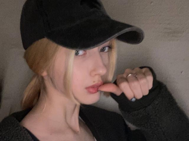 Primo piano di MarcellaMcleskey, una donna bionda con la pelle chiara, che indossa un cappello nero e una maglietta nera, mentre succhia una carota. Ha gli occhi azzurri e un'espressione seria. L'illuminazione è fioca..