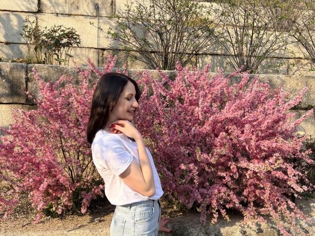 Fredericka Thonney, con cabello oscuro de longitud media, una camiseta blanca y jeans de cintura alta, de pie junto a un color rosa vibrante. arbusto en flor. Ella está tocándose el pecho, mirando hacia un lado con una sonrisa sutil. Iluminado por el sol.