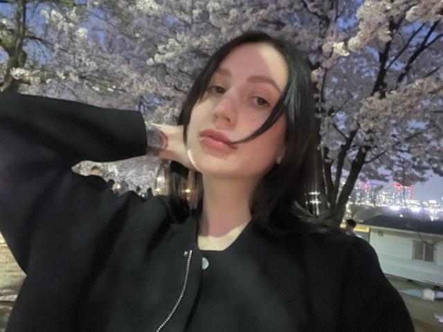 Photographie de Fredericka Thonney, le visage légèrement incliné, cheveux noirs, portant une chemise noire, la main derrière la tête, fleurs de cerisier. arbres en arrière-plan, lumières de la ville visibles, séduisant, confiant, pose décontractée, lumière du soir..