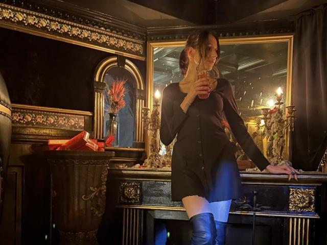 Fredericka Thonney steht in einem prunkvollen, dunklen Raum mit aufwendigen Goldakzenten, hält ein Telefon, trägt ein schwarzes Kleid und blaue, hohe Strumpfhosen, die bis zum Oberschenkel reichen. Kerzen beleuchten ihre sinnliche, verführerische Pose..