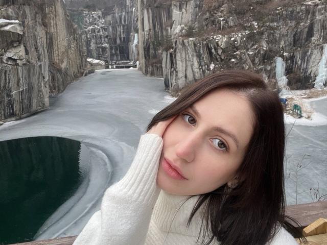 Fredericka Thonney, una mujer de piel clara con cabello largo y negro, vestida con un cuello alto blanco, posa frente a un paisaje congelado. Cañón rocoso con hielo cubriendo el suelo. Ella toca su mejilla, mirando hacia adelante.