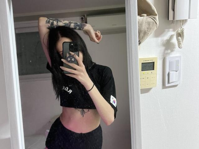 Fredericka Thonney, mit schwarzen Haaren, tätowierter Arm angehoben, schwarzes Crop-Top, schwarze Hose, Spiegelbild, Handy in der Hand, sichtbar. Bauch-Tattoo, helles Badezimmer, weiße Wände, Ventilator, Lichtschalter, lässig, sexy, selbstbewusste Pose..