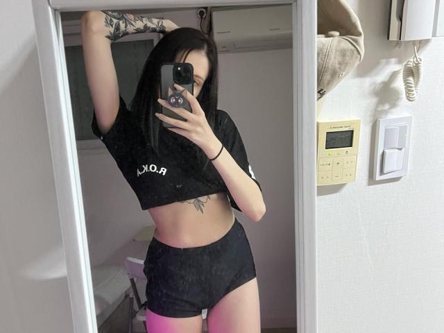 Fredericka Thonney maakt een spiegelzelfportret, waarop haar slanke, getatoeëerde lichaam te zien is in een zwart topje en korte broek. Ze houdt haar telefoon met één hand vast, waardoor haar platte buik en suggestieve kleding zichtbaar zijn. Helder licht.