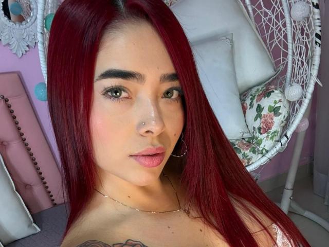 Nahaufnahme von ValeriaRoseT mit langen, glatten roten Haaren, heller Haut und einem Nasenpiercing. Sie blickt direkt in die Kamera, ihre Lippen sind zu einem Schmollmund geformt, und sie trägt eine Halskette. Stuhl mit Blumenmuster im Hintergrund.