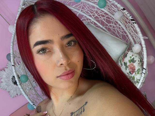Foto em close-up de ValeriaRoseT com cabelo longo e liso, ruivo, pele clara e olhos castanhos. Ela tem um piercing no nariz, uma tatuagem na clavícula e uma expressão neutra. Cadeira de macramé branca com bolinhas em tons de verde-água e branco..