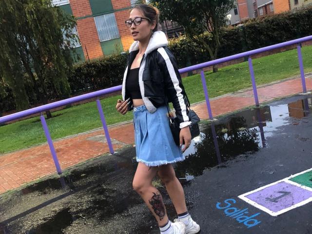 Zoe Taylorl, avec sa peau foncée et ses lunettes, marche sur un trottoir mouillé, vêtue d'une jupe bleue et d'un sweat à capuche noir, mettant en valeur un grand tatouage sur sa cuisse droite. Elle porte des baskets blanches..