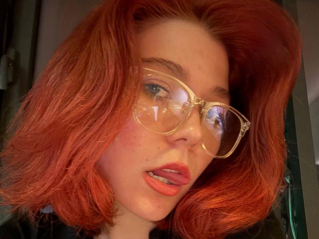 Fotografía de primer plano de GerdaCastin con cabello rojo y ondulado, usando gafas grandes con montura dorada y pintalabios rojo. Tiene una expresión sensual, con los labios ligeramente entreabiertos. Su piel es clara y tiene algunas pecas..