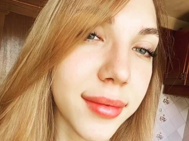 Close-up do rosto de Alease Benson, com cabelo castanho claro, olhos azuis e lábios vermelhos. A luz do sol realça sua pele lisa. Ela está sorrindo discretamente, criando uma sensação de tensão sexual sutil..