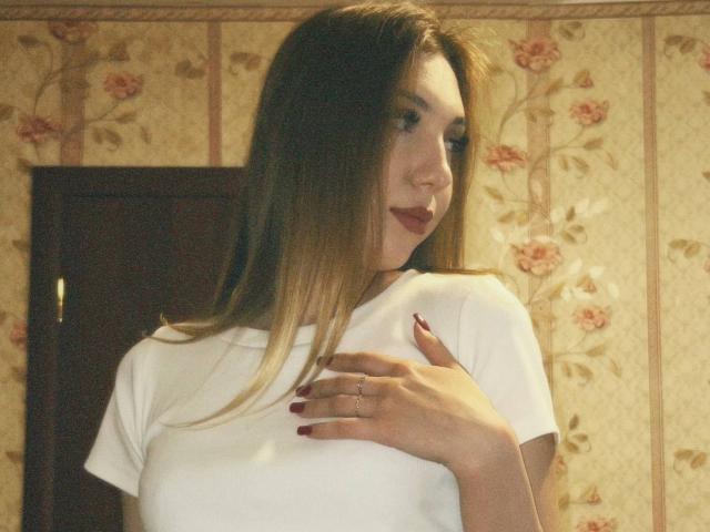 Alease Benson está em um quarto com papel de parede floral, vestindo uma camiseta branca, cobrindo o peito com uma mão, aparentemente cabelo castanho, batom vermelho e unhas de vermelho escuro. Tensão sexual evidente..