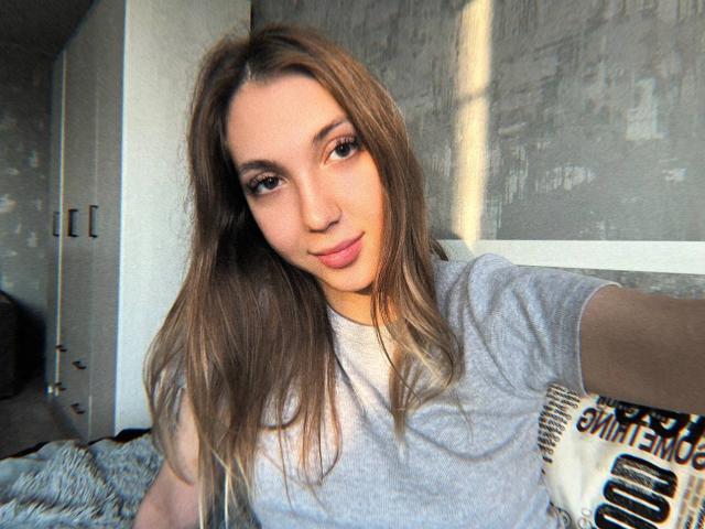 Alease Benson, com cabelos castanhos longos, vestindo uma camiseta cinza, segurando um violão, olhando para a câmera com um leve sorriso, luz natural e brilhante, ambiente moderno, selfie casual, atmosfera relaxada..