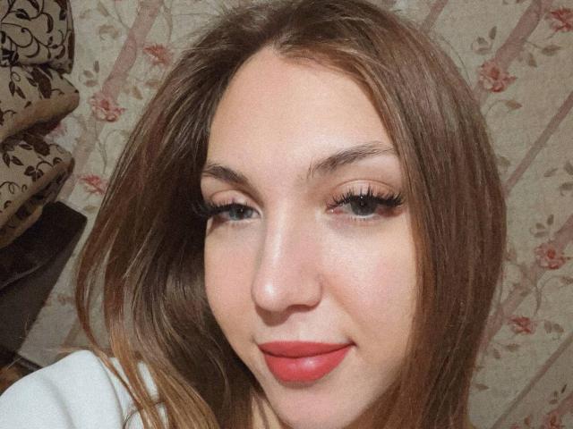 Close-up van Alease Benson, een vrouw met een lichte huid, lang bruin haar, rode lippenstift en veel eyeliner. Ze glimlacht lichtjes en toont een vleugje seksuele spanning door direct oogcontact te maken..