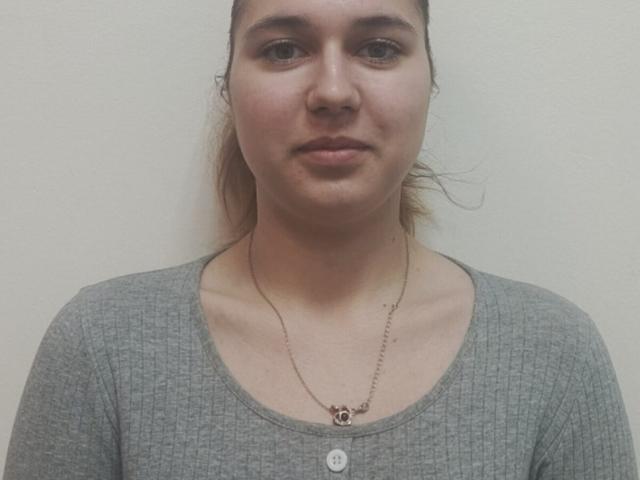 Nahaufnahme von JenaWylde mit einem neutralen Gesichtsausdruck, heller Haut, braunen Augen und braunem Haar, das zu einem Dutt gebunden ist. Sie trägt ein graues, geripptes Hemd und eine feine Halskette mit einem kleinen Anhänger..