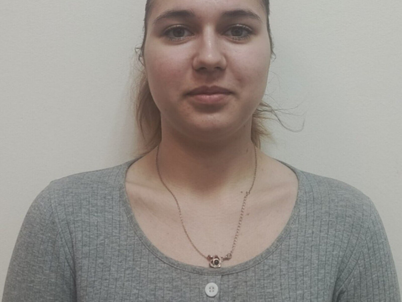 Gros plan de JenaWylde, avec une expression neutre, une peau claire, des yeux marron et des cheveux bruns attachés en arrière. Portant une chemise gris côtelée et un collier fin avec un petit pendentif..