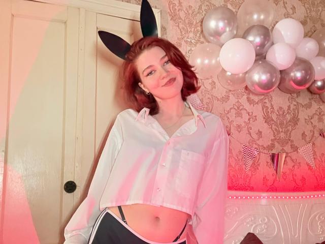 GerdaCastin, red-haired woman with black bunny ears, wearing a white crop top and black shorts, standing in a room with silver balloons and patterned wallpaper, posing seductively. Bright red lighting.