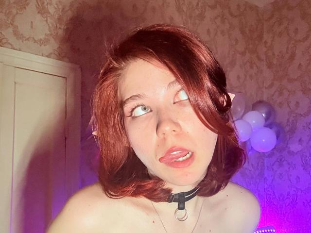 Photo numérique de Gerda Castin avec une coupe de cheveux courte et rouge, peau claire, yeux bleus, la langue tirée, portant un collier noir, de couleur violet clair. et un arrière-plan avec un effet bokeh rose, tension sexuelle, éclairage vif, expression suggestive..