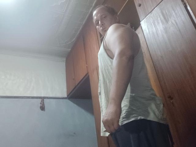 Fotografía de maxKnd-hot, un hombre musculoso con la cabeza calva, que lleva una camiseta blanca y pantalones negros, de pie en un lugar tenuemente iluminado. Habitación iluminada con armarios de madera y una pared blanca. Está mirando hacia atrás con una expresión seria. Increíblemente intenso..