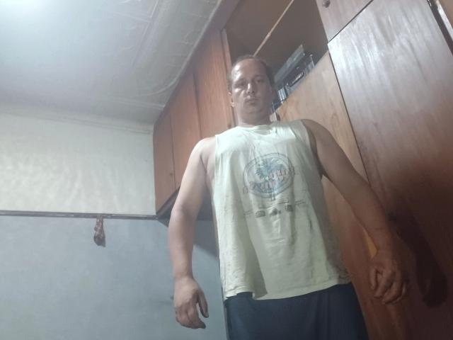 Photograph of maxKnd-hot standing in a room, wearing a sleeveless white shirt with a graphic, black pants. He's looking down, arms relaxed, wood cabinets in the background. Fucking tense vibe.