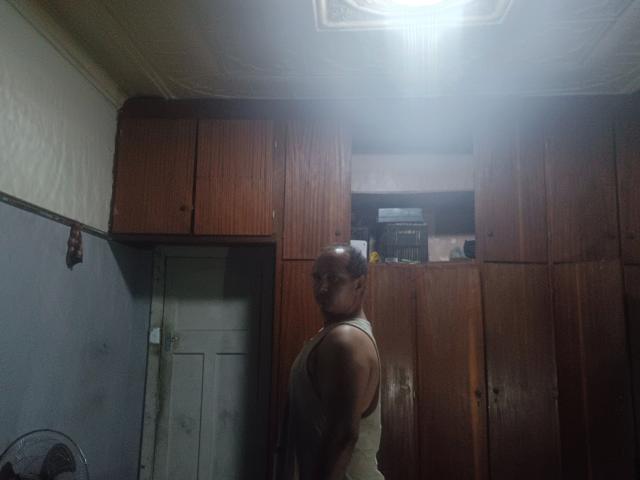 Fotografia di un uomo muscoloso, calvo, con la pelle abbronzata, in piedi in una stanza poco illuminata, che indossa una maglietta senza maniche e guarda verso l'obiettivo. lontano, sotto una luce intensa a soffitto, armadi in legno sullo sfondo.