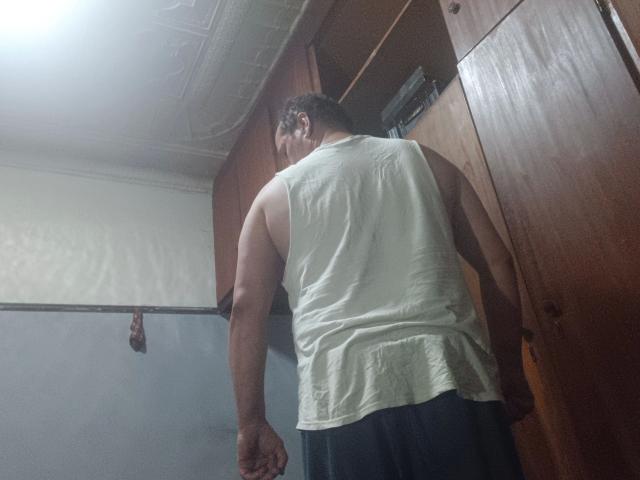 Photograph of maxKnd-hot, muscular man with tan skin, white sleeveless shirt, black pants, leaning forward, showing defined back muscles and a bulge in his pants, in a dimly lit room.