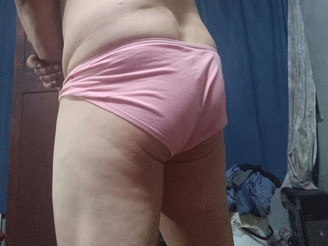 Close-up foto van de onderrug en billen van een aantrekkelijke persoon, gekleed in strakke, roze onderbroek. Hun hand rust op hun heup, waardoor een lichte kromming in hun onderrug zichtbaar is. Kleding op de vloer.