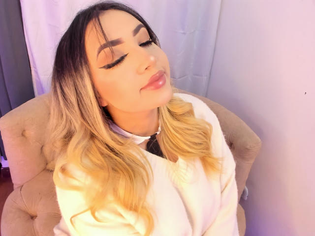 MelissaLust, a woman with long blonde and black hair, eyes closed, wearing a white shirt, sitting in a beige tufted chair, sunlight casting a bright glow on her face, lips slightly parted.