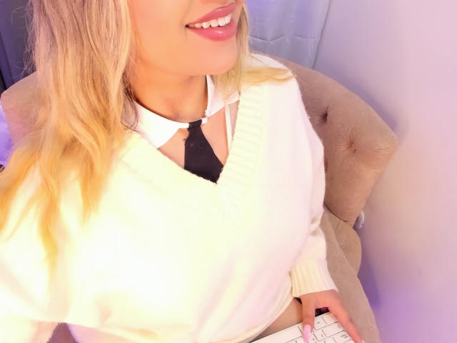 Close-up photo of MelissaLust, blonde hair, white shirt, black tie, smiling with pink lips, hand on beige tufted chair, visible white keyboard, bright light, sexual tension, provocative pose.