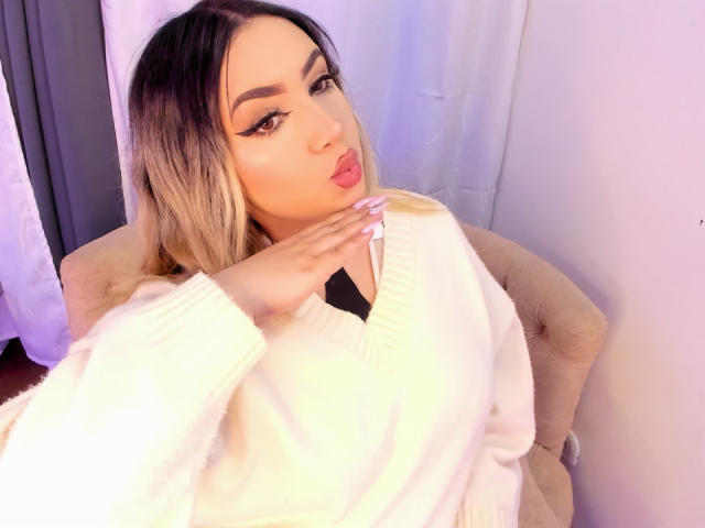 MelissaLust, a sexy woman with long, ombre hair, gazes up with pouty lips. She wears a fluffy white sweater, touching her chest with a seductive look. Bright, white background.