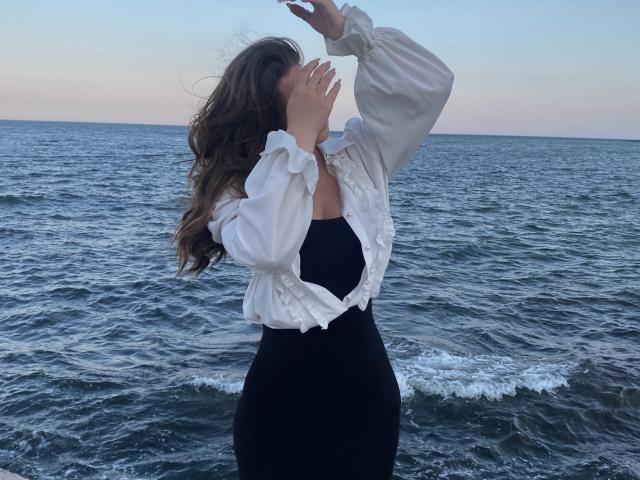 Miss Kuromi está até a cintura na água do oceano, com os braços levantados e a cabeça inclinada para trás. Ela veste uma blusa branca com babados e uma saia preta, enquanto o vento sopra em seu longo cabelo castanho. Muito atraente..