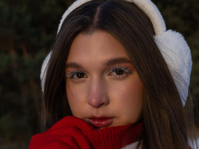 Close-up photo of MissKuromi with dark brown hair, wearing a white hat and red scarf, gazing intensely with sparkling eyes. Her face is close to the camera, creating a sexual tension vibe.