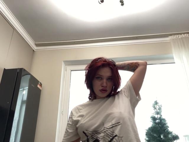 PeggyBehimer, red-haired, standing in a room, white t-shirt with black design, arm raised behind head, intense expression, natural light from window, black cabinet on left, tree visible outside.