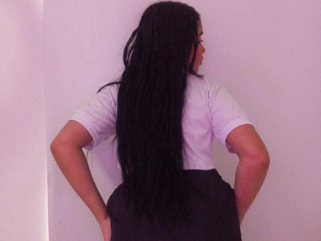 AliceSmity, with long black hair, stands with hands on hips, facing away, wearing a white t-shirt and black skirt. Her posture exudes confidence and a hint of sexual tension.