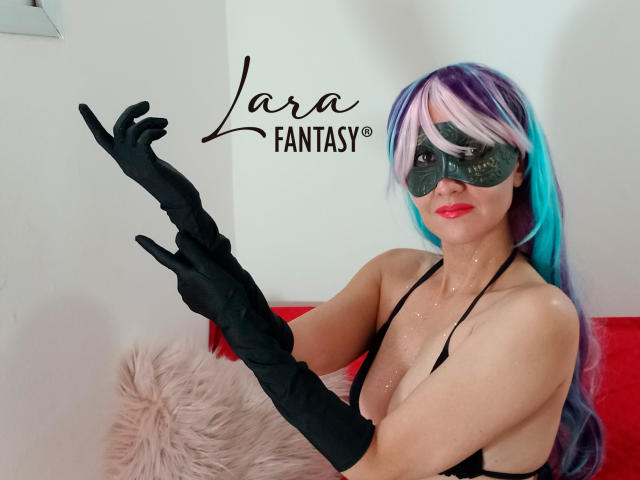 LaraFantasy, usando uma máscara preta e luvas pretas longas, tem cabelo azul e branco, lábios vermelhos e um top preto de alças. gesticulando com a mão direita. Texto diz "Lara FANTASIA"."