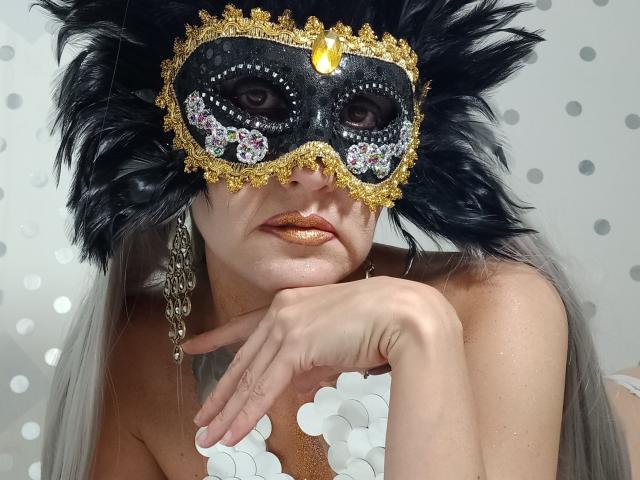 Foto em close-up de LaraFantasy usando uma máscara de carnaval preta e dourada com detalhes de penas, e glitter dourado ao redor. olhos, e cabelos longos e grisalhos. Ela apoia o queixo na mão, com um olhar sedutor..