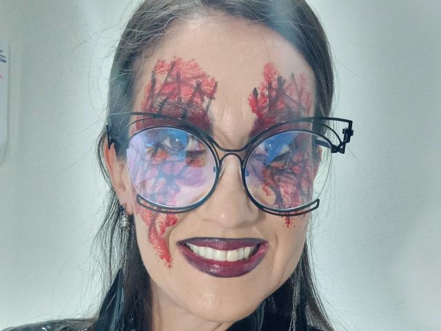 Close-up photo of LaraFantasy with smeared red paint on her face, wearing reflective black-rimmed glasses, and dark red lipstick. She's smiling with visible white teeth. Background is plain white.
