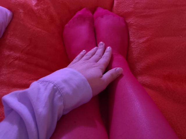 Gros plan des pattes rouges de Marriam, croisées sur une surface rouge, avec une main gantée de blanc reposant sur ses pieds. Des teintes rouges vives, un contact intime, une tension érotique subtile, une pose décontractée..