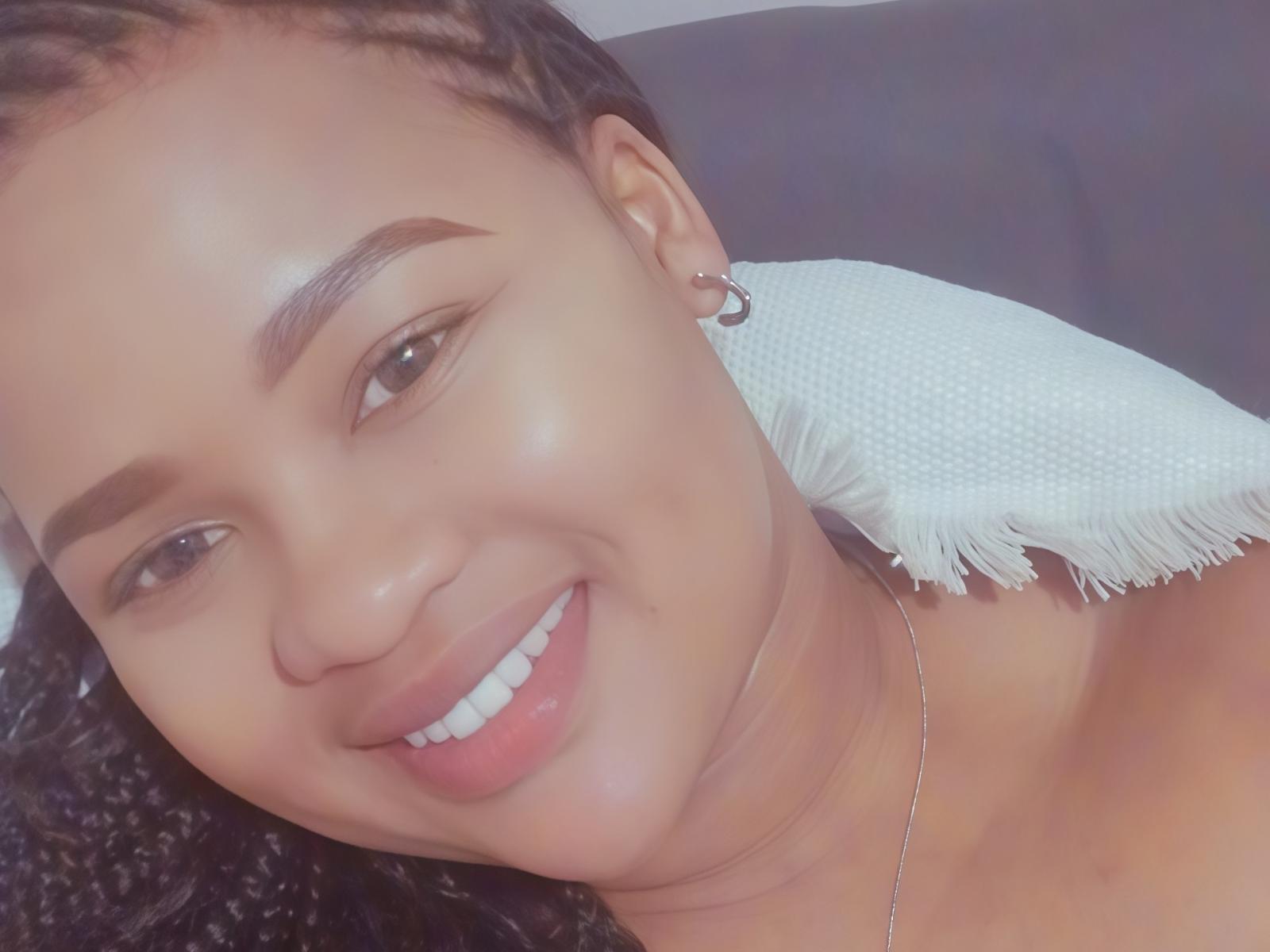 Selfie de primer plano de Devinesa, con una cara sonriente, piel morena y cabello negro rizado. Ella lleva un pequeño aro en la oreja y un collar de plata, sobre una superficie blanca con flecos..