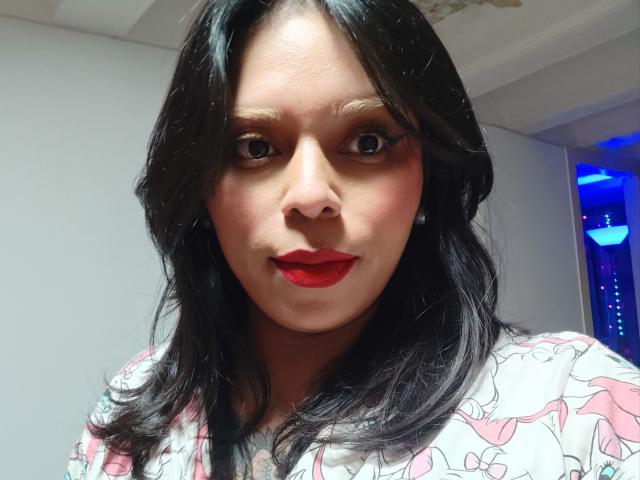 Close-up photo of Chili with medium brown skin, black wavy hair, red lipstick, and a white floral shirt. She looks directly at the camera with a slight smile, exuding sexual tension.