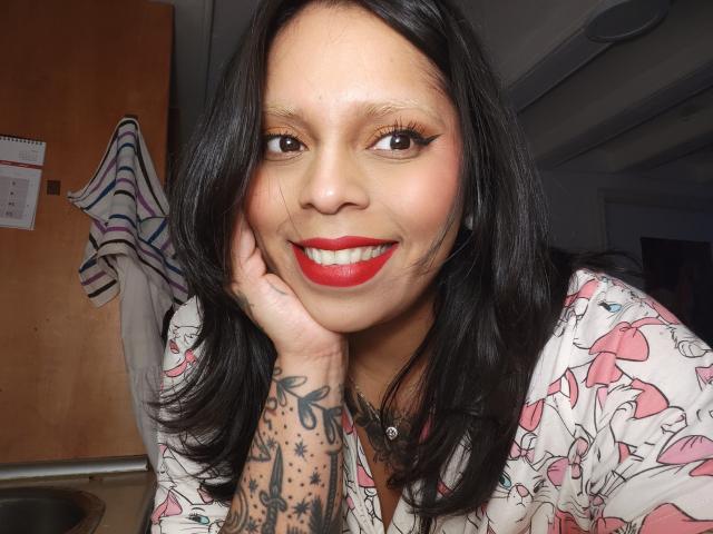 Close-up of Chili with medium brown skin, black hair, red lipstick, and a tattooed hand resting on her cheek. She's smiling with a hint of sexual tension, wearing a floral shirt.