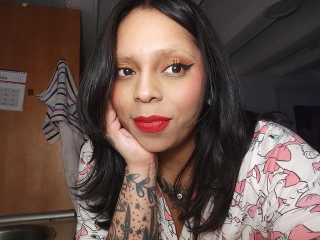 Close-up of Chili, an Asian woman with black hair, red lipstick, and tattoos on her right hand. She rests her head on her hand, looking directly at the camera with seductive eyes. She's wearing a white floral shirt. Her red lipstick contrasts with her pale skin. Bright lighting highlights her face.