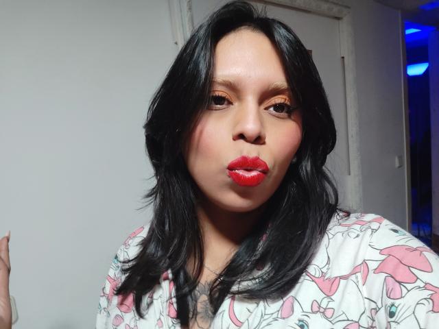Photograph of a plus-sized woman with dark, wavy hair, red lipstick, and a white floral shirt, pouting her lips. Bright lighting highlights her face, creating a sexual tension vibe.